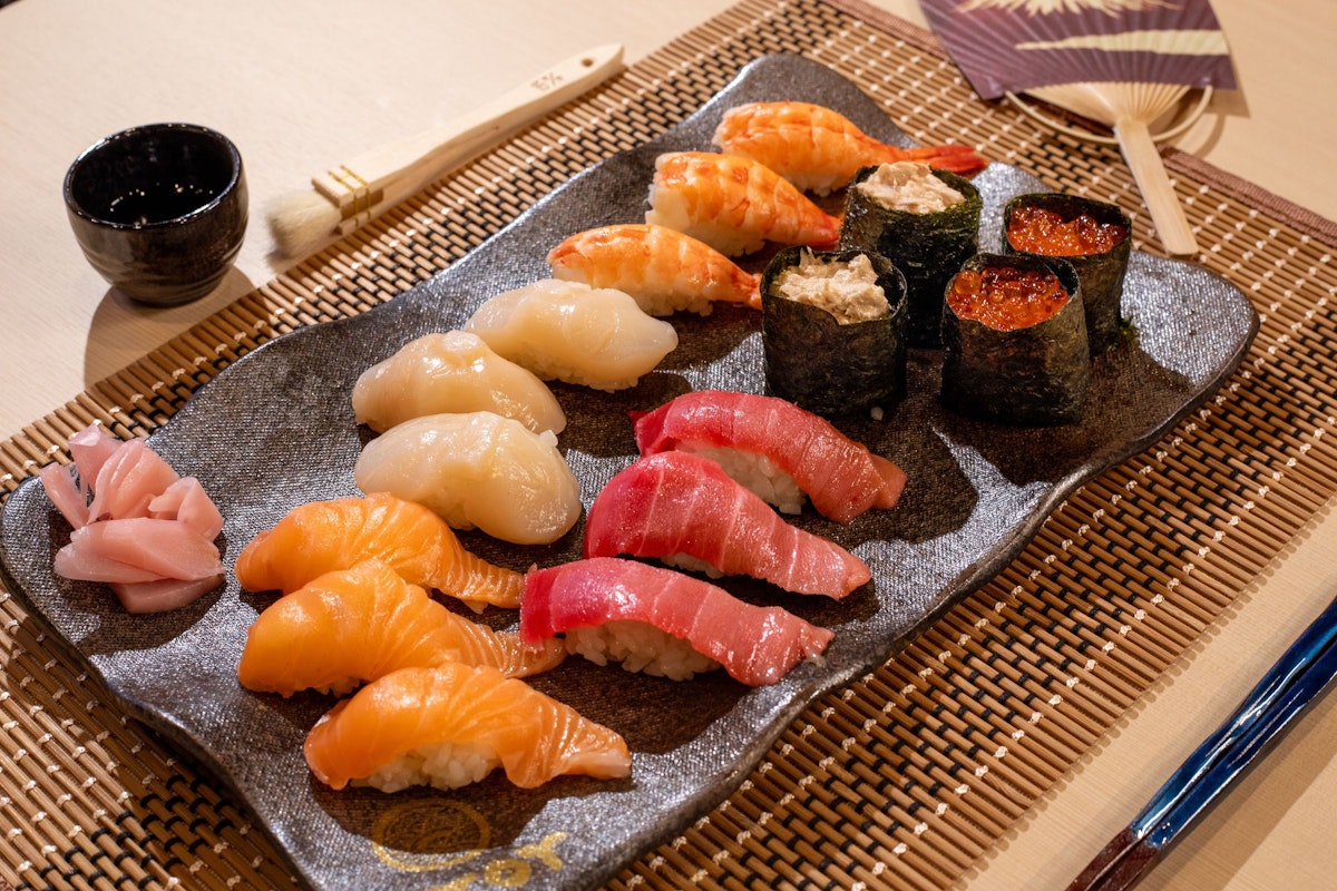 Sushi Making Experience A plate of assorted sushi on a bamboo placemat, featuring salmon, tuna, scallops, shrimp, and roe-topped rolls. Served with pickled ginger, soy sauce in a small cup, and a pair of chopsticks. An open fan is placed beside the plate.