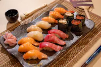 A plate of assorted sushi on a bamboo placemat, featuring salmon, tuna, scallops, shrimp, and roe-topped rolls. Served with pickled ginger, soy sauce in a small cup, and a pair of chopsticks. An open fan is placed beside the plate.