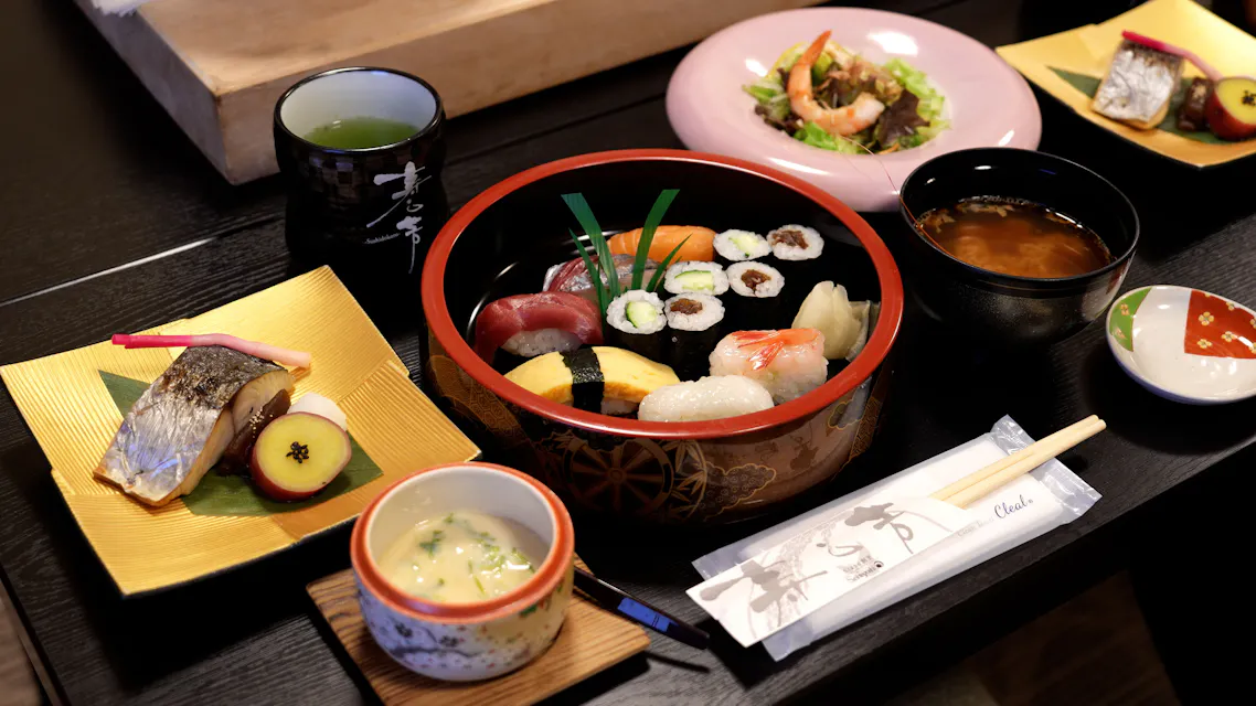 An assortment of Japanese dishes includes sushi rolls and nigiri in a bowl, grilled fish, chawanmushi (steamed egg custard), miso soup, and tempura on separate plates. Chopsticks and a ceramic cup complete the setting on a dark table.