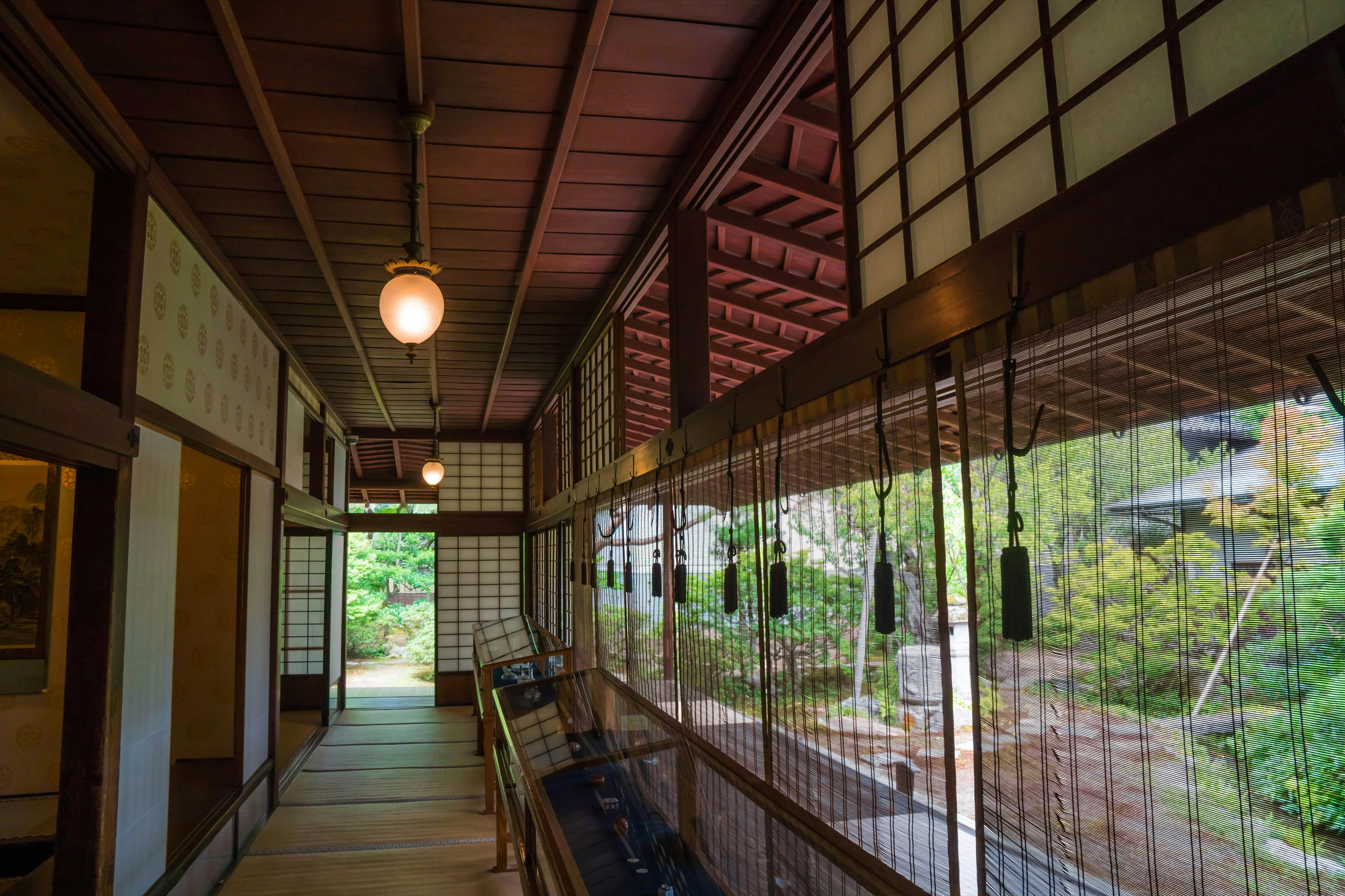 A traditional Japanese wooden hallway with tatami mats, shoji screens, and hanging light fixtures, overlooking a garden with lush greenery through large windows and bamboo blinds.
