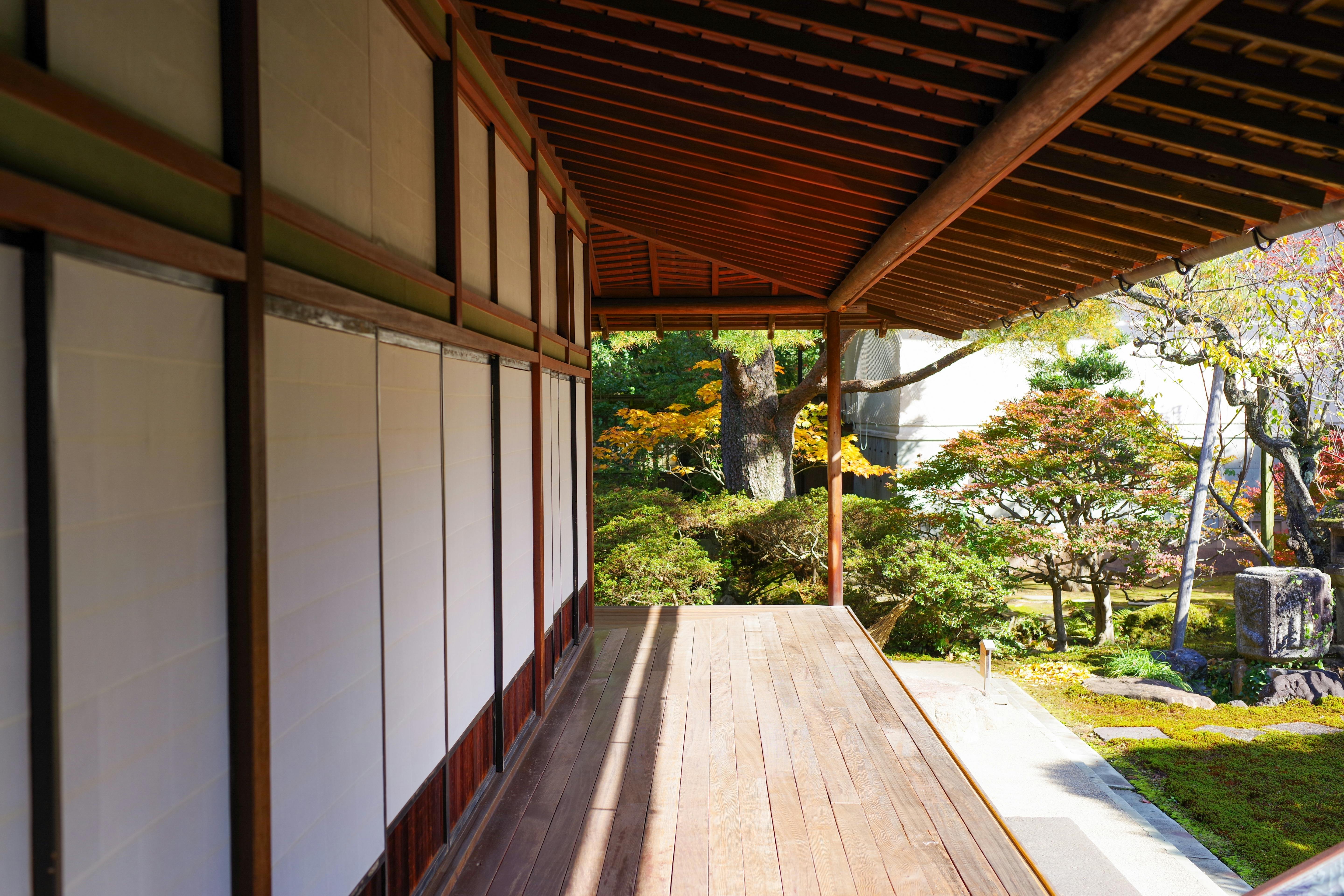 A wooden veranda extends along the side of a traditional Japanese building, overlooking a tranquil garden with green trees, shrubs, and sunlight filtering through the foliage.