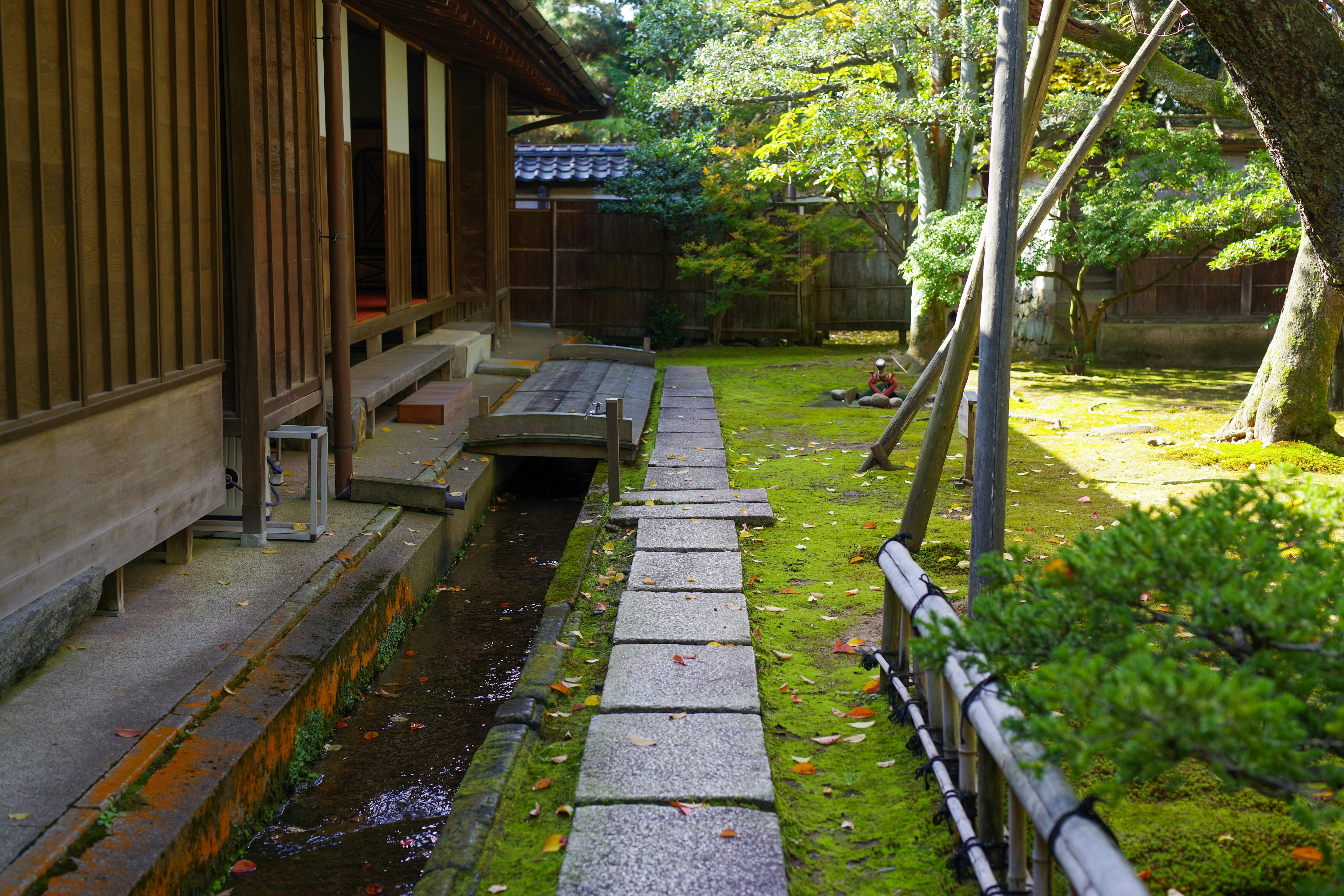 A peaceful Japanese garden with a stone path, moss-covered ground, a wooden building, and trees casting dappled sunlight.