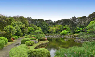 A peaceful Japanese garden with manicured bushes, a winding stone path, and a tranquil pond surrounded by lush green trees under a clear blue sky.