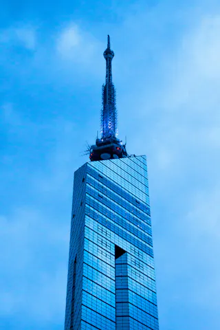 The image shows the top portion of a modern skyscraper with reflective blue-tinted glass windows and a tall spire against a cloudy sky. The building's sharp lines and antenna are prominent.