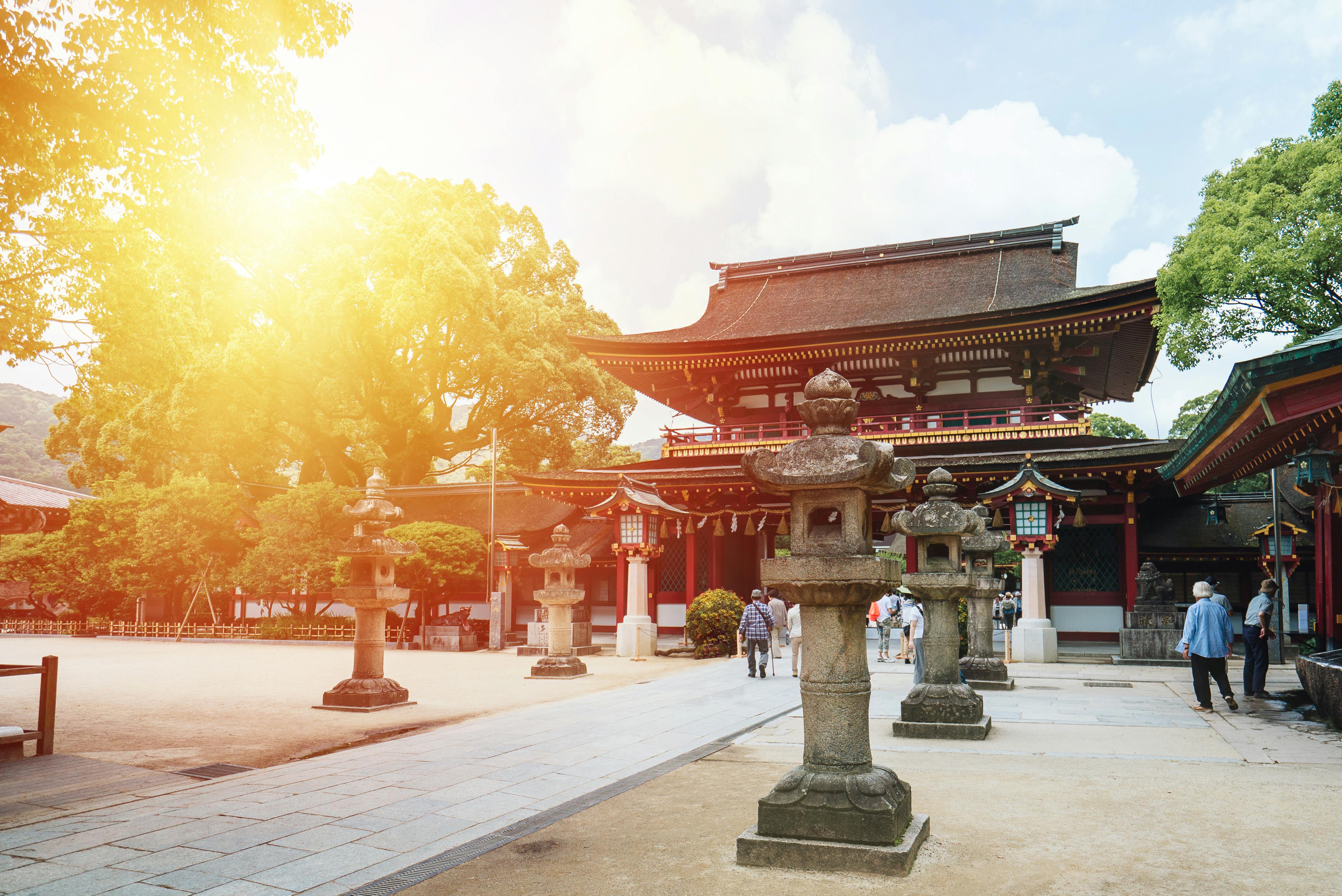 A traditional Japanese shrine with stone lanterns, red wooden pillars, and a curved roof, surrounded by trees and visitors walking in the sunlight.