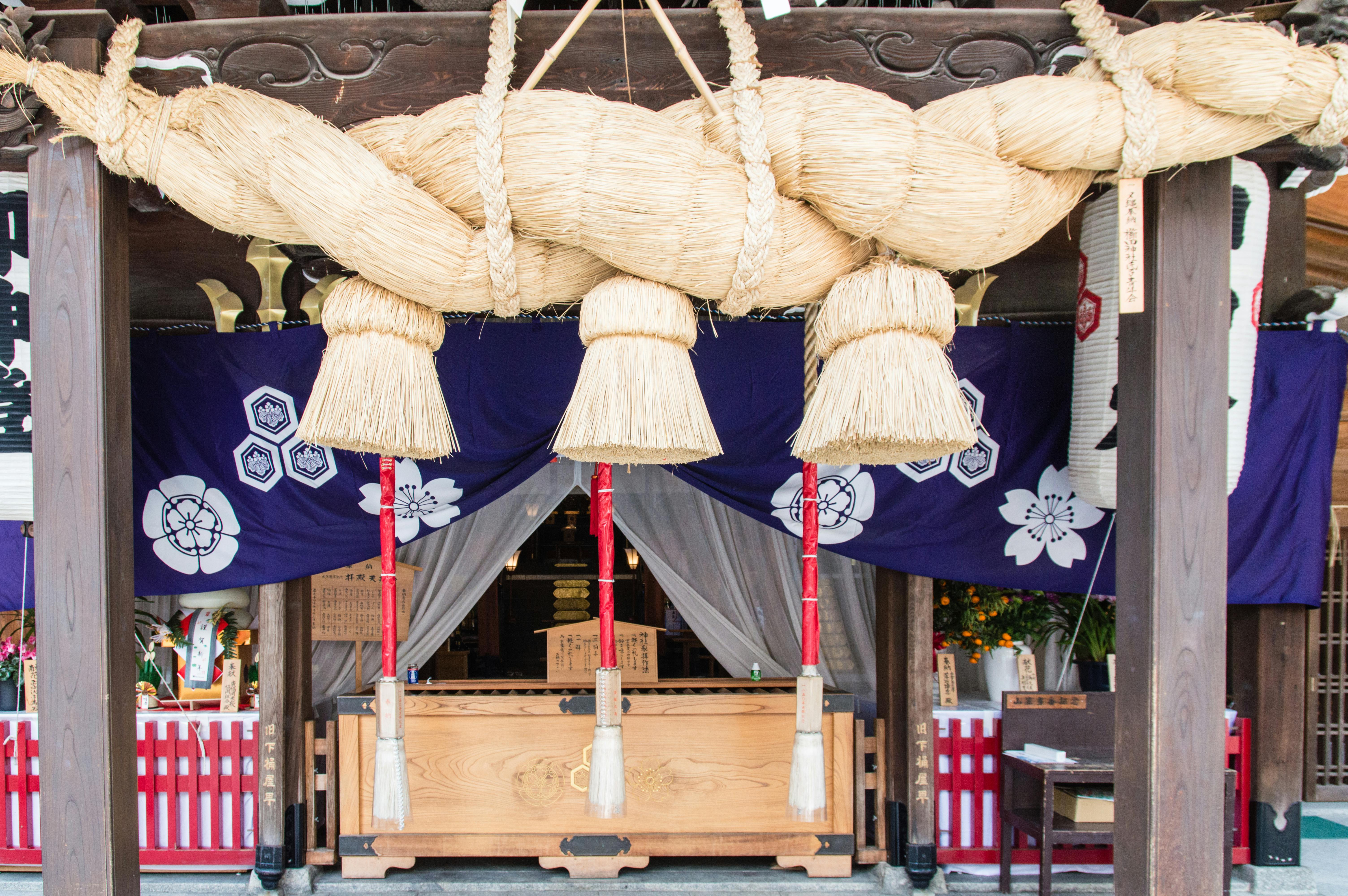 Large braided straw rope with hanging tassels displayed at the entrance of a Japanese shrine, with purple curtains, wooden pillars, and a traditional offering box underneath.