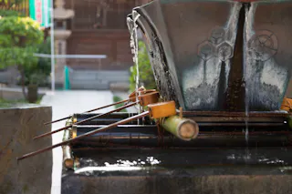 A traditional Japanese purification fountain with wooden ladles resting on bamboo poles, water gently flowing from a metal spout into a stone basin, set in an outdoor temple area.