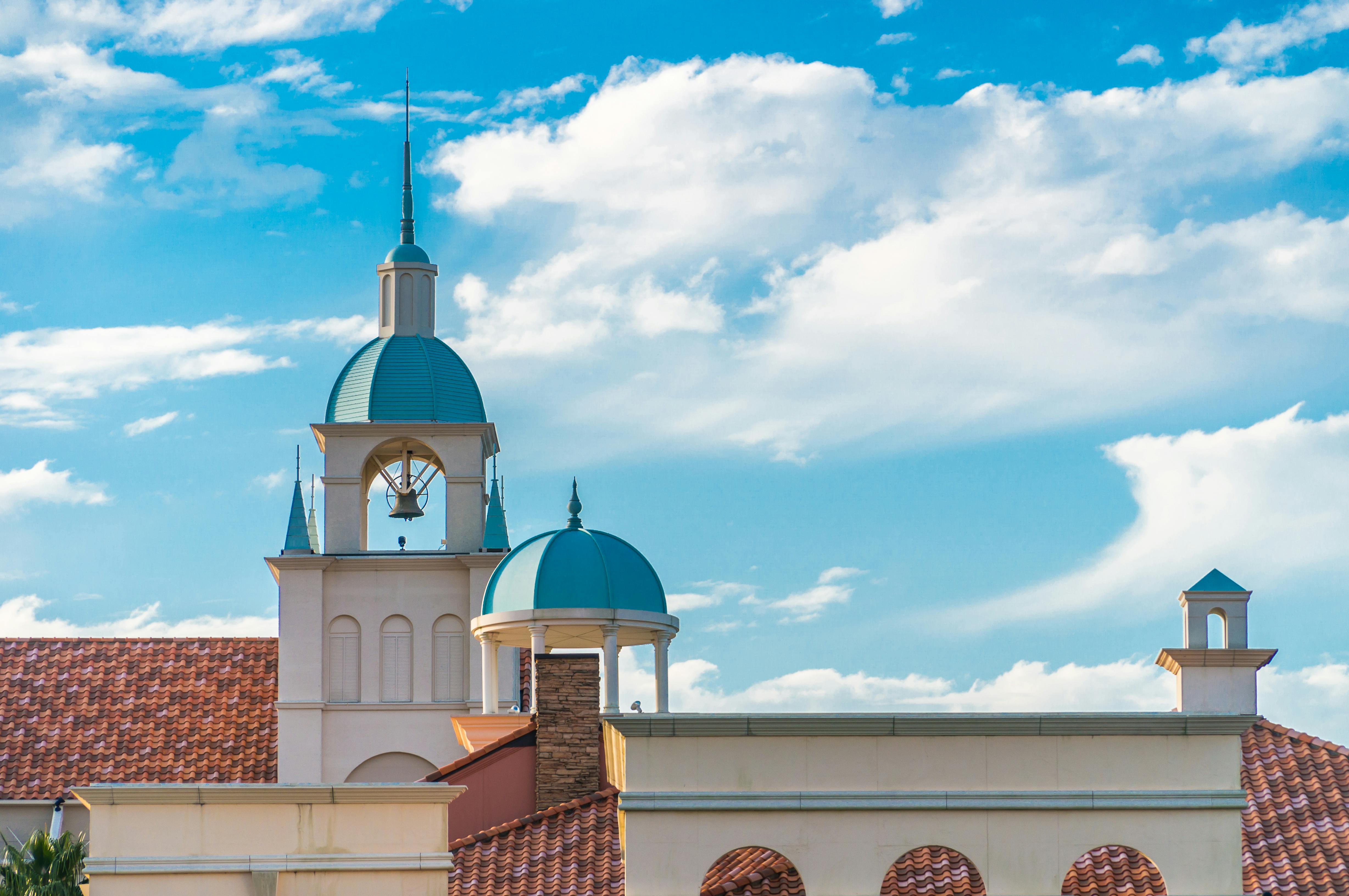 A building with a red-tiled roof and white walls features a central clock tower and smaller domes, all with blue-green roofs, under a bright sky with scattered white clouds.