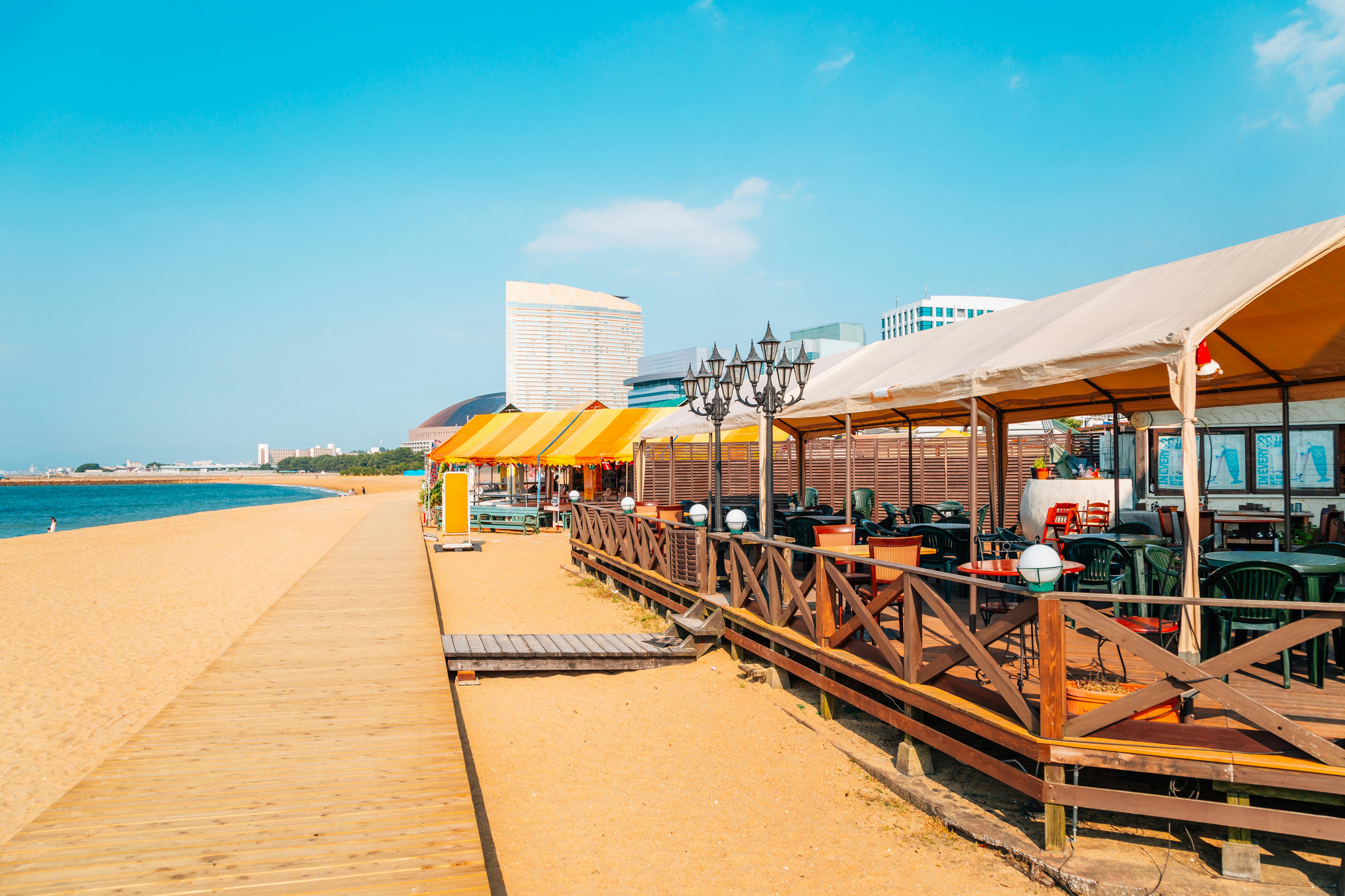 A sunny beach boardwalk with outdoor restaurants under tents, wooden railings, and empty tables and chairs. Tall city buildings are in the background, and the blue sky and ocean are visible on the left.