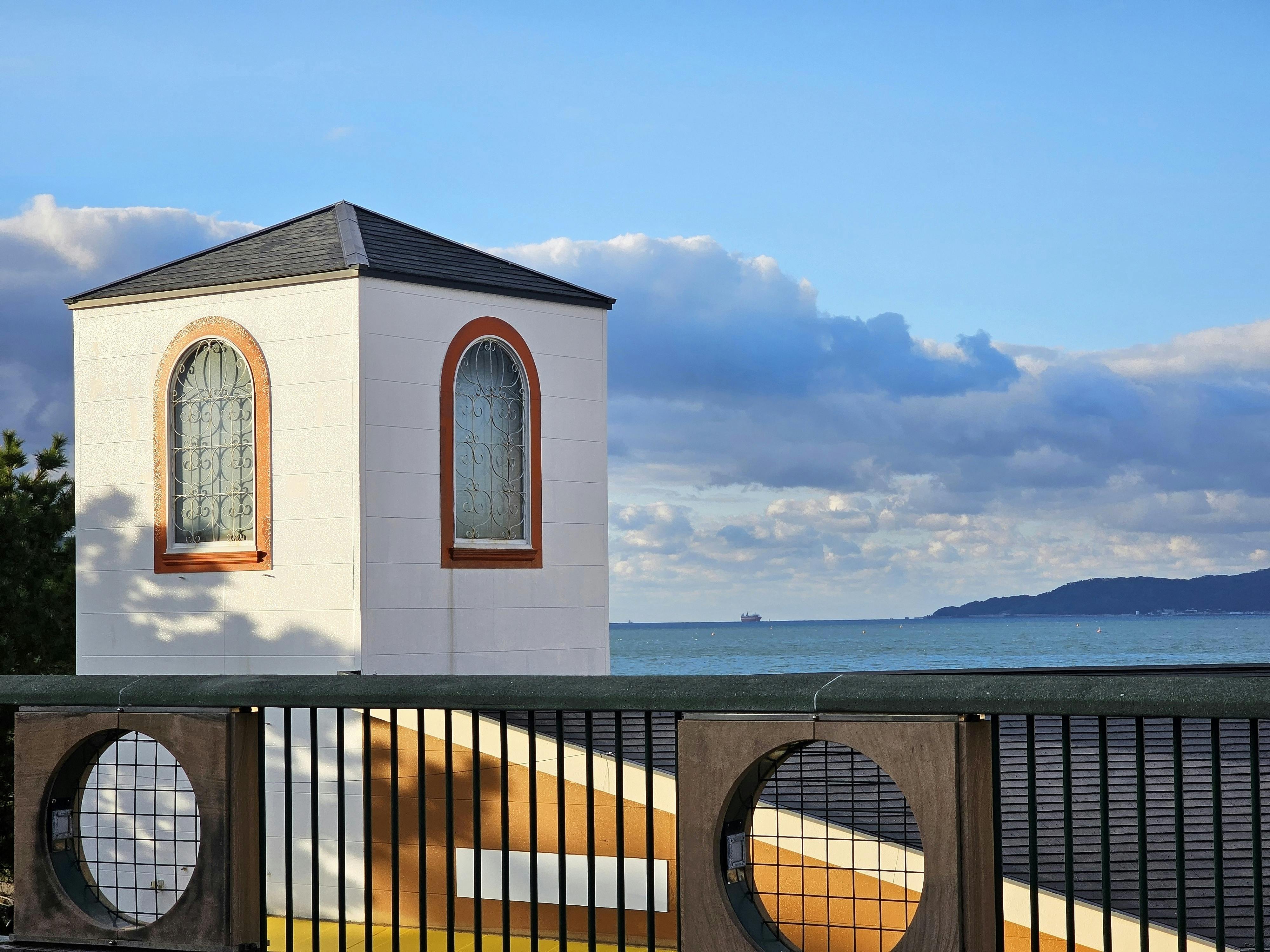 A small white building with arched windows stands near a black fence with circular designs, overlooking the ocean and distant hills under a partly cloudy sky.