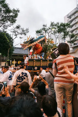 A crowd watches a colorful festival float decorated with a large dragon and lanterns being paraded through a street lined with trees and buildings. Some people are taking photos, while others observe or sit on shoulders for a better view.