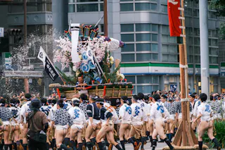 A large group of people in traditional Japanese clothing carry a decorated float with cherry blossoms through city streets during a festival. Water splashes in the air, and modern buildings are visible in the background.