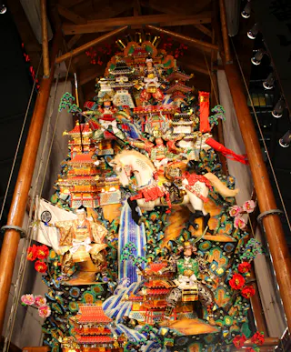A tall, colorful Japanese festival float decorated with elaborate figures, a white horse, warriors in armor, vibrant buildings, cascading blue water, and red flowers, set under a wooden roof.