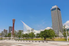 Modern cityscape featuring a tall skyscraper, a red observation tower, and a unique white, sail-like structure with trees and a paved plaza in the foreground under a clear blue sky.