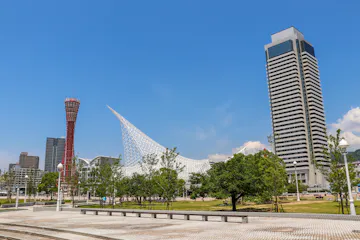 Modern cityscape featuring a tall skyscraper, a red observation tower, and a unique white, sail-like structure with trees and a paved plaza in the foreground under a clear blue sky.