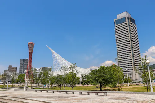 Modern cityscape featuring a tall skyscraper, a red observation tower, and a unique white, sail-like structure with trees and a paved plaza in the foreground under a clear blue sky.