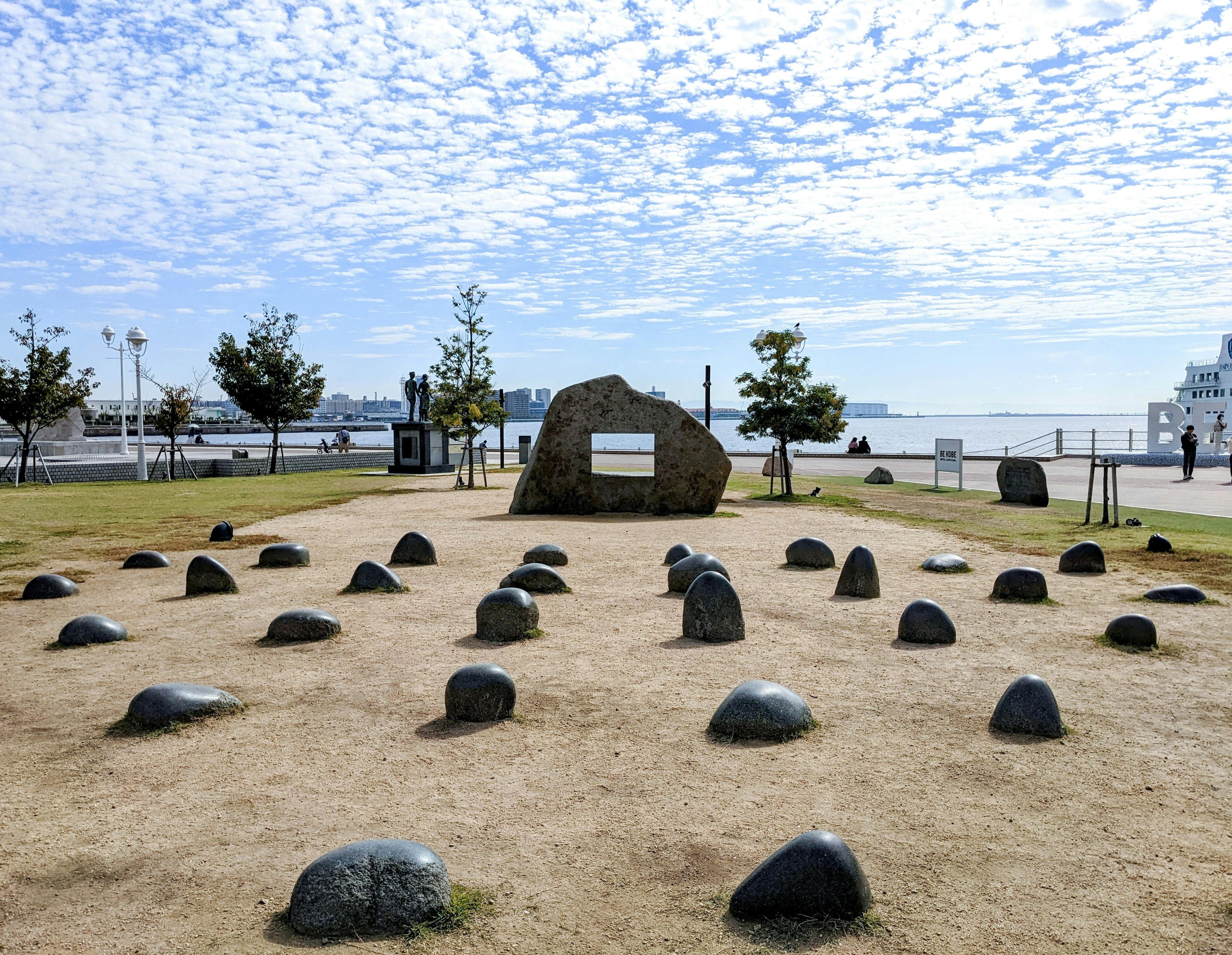 A park area with numerous black stones arranged on sandy ground, a large central rock with plaques, some trees, and a waterfront view under a partly cloudy sky.