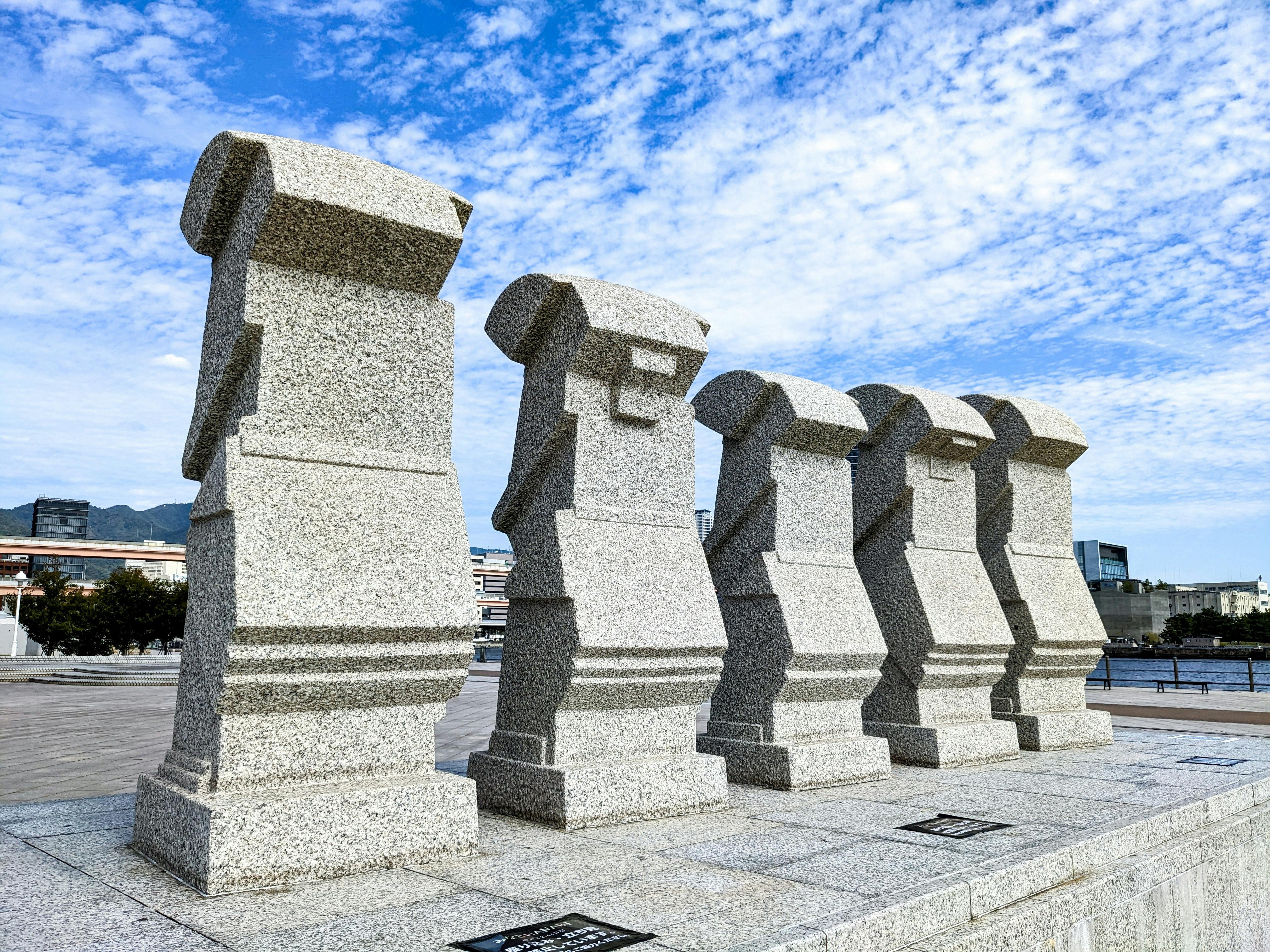 Five large stone statues resembling Moai heads stand in a row on a paved plaza under a partly cloudy sky, with city buildings and trees visible in the background.