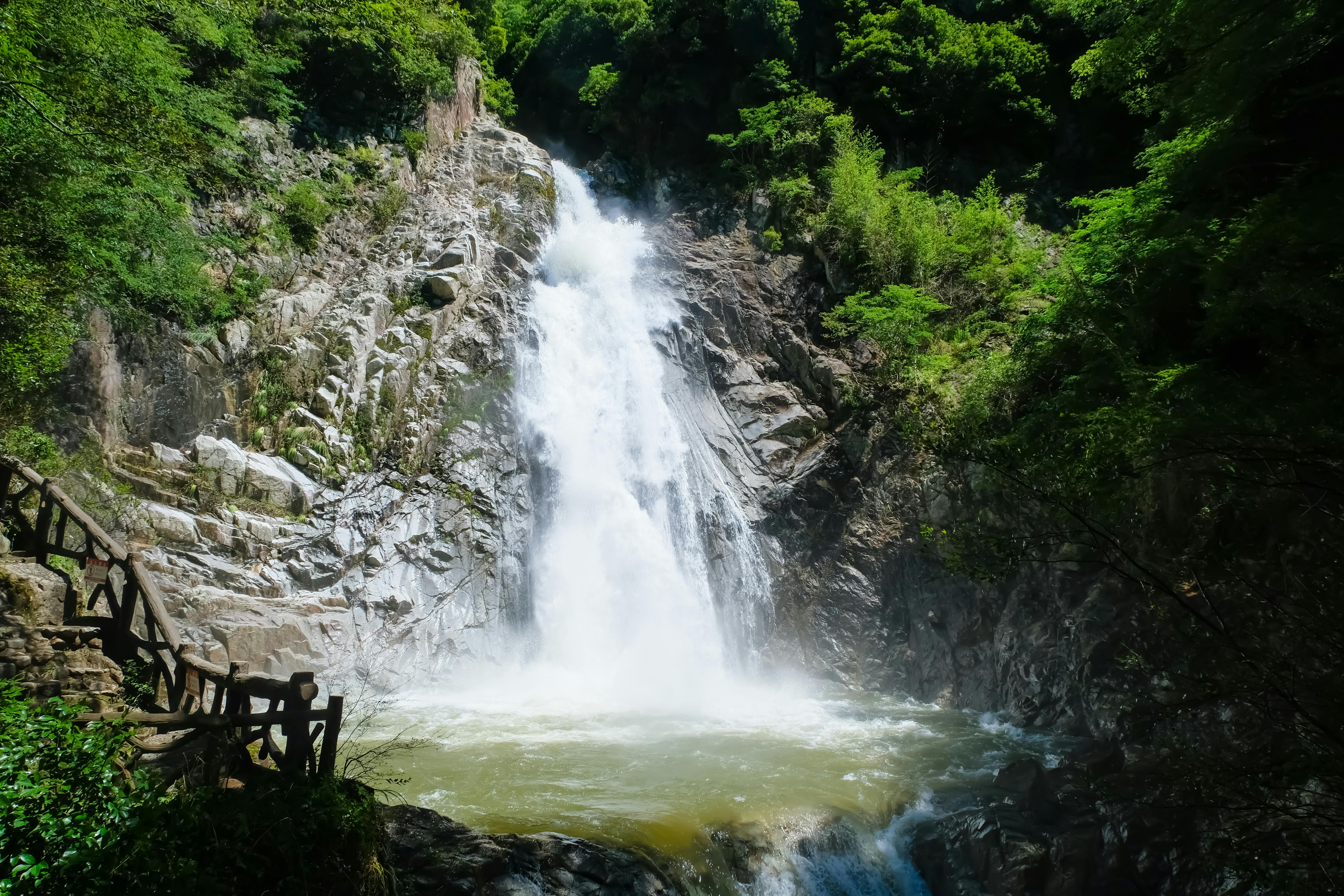 A powerful waterfall cascades down rocky cliffs into a pool surrounded by lush green trees, with a wooden railing path visible on the left side.