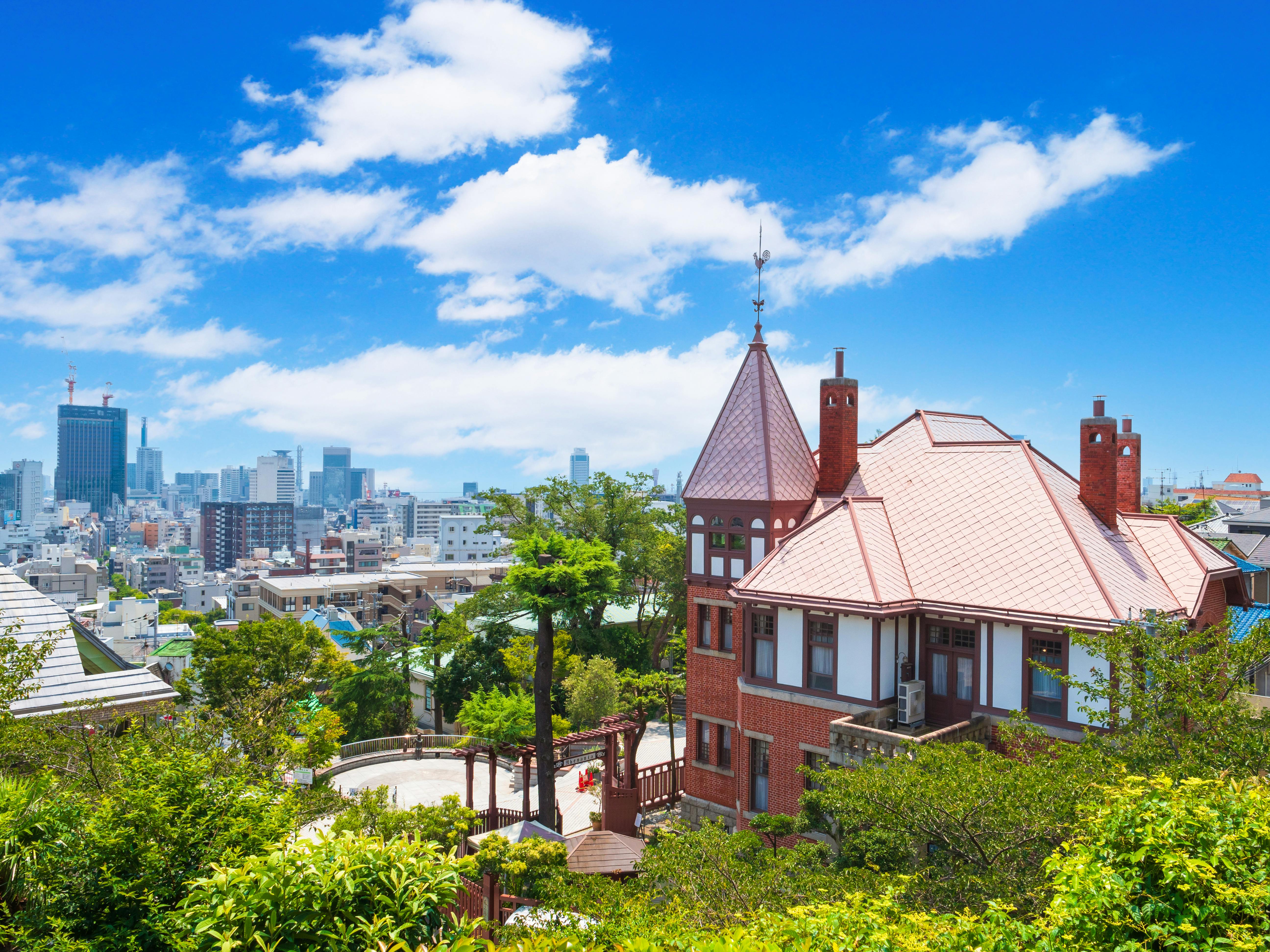 A red-brick, Western-style house with a pointed tower sits among green trees, overlooking a modern city skyline under a bright blue sky with scattered clouds.
