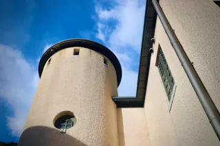 A low-angle view of a beige, textured building with a round tower and rectangular windows, set against a blue sky with scattered clouds.