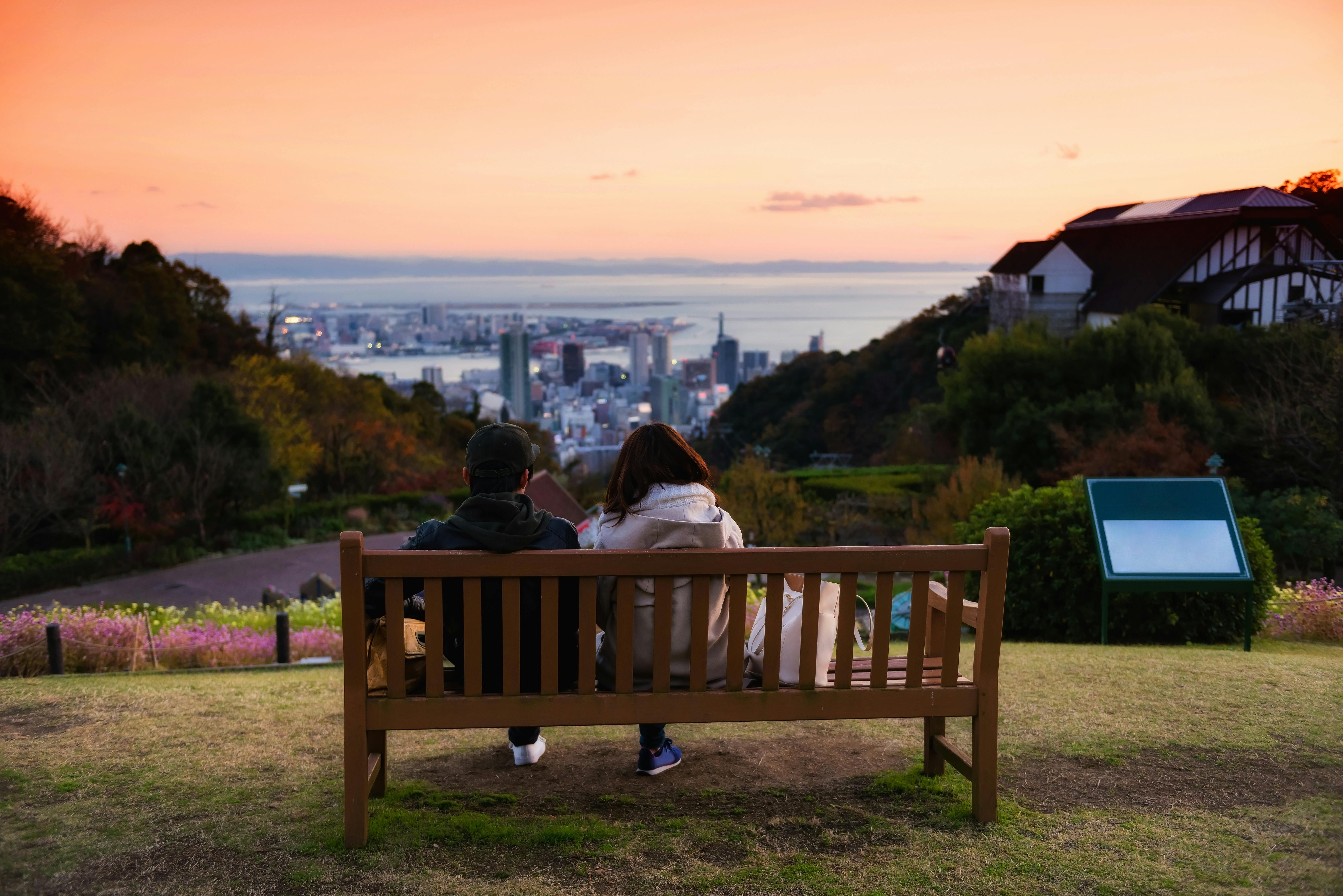 A couple sits on a wooden bench overlooking a cityscape at sunset, with vibrant orange skies, buildings below, and greenery surrounding them. A scenic viewpoint sign stands nearby on the grass.