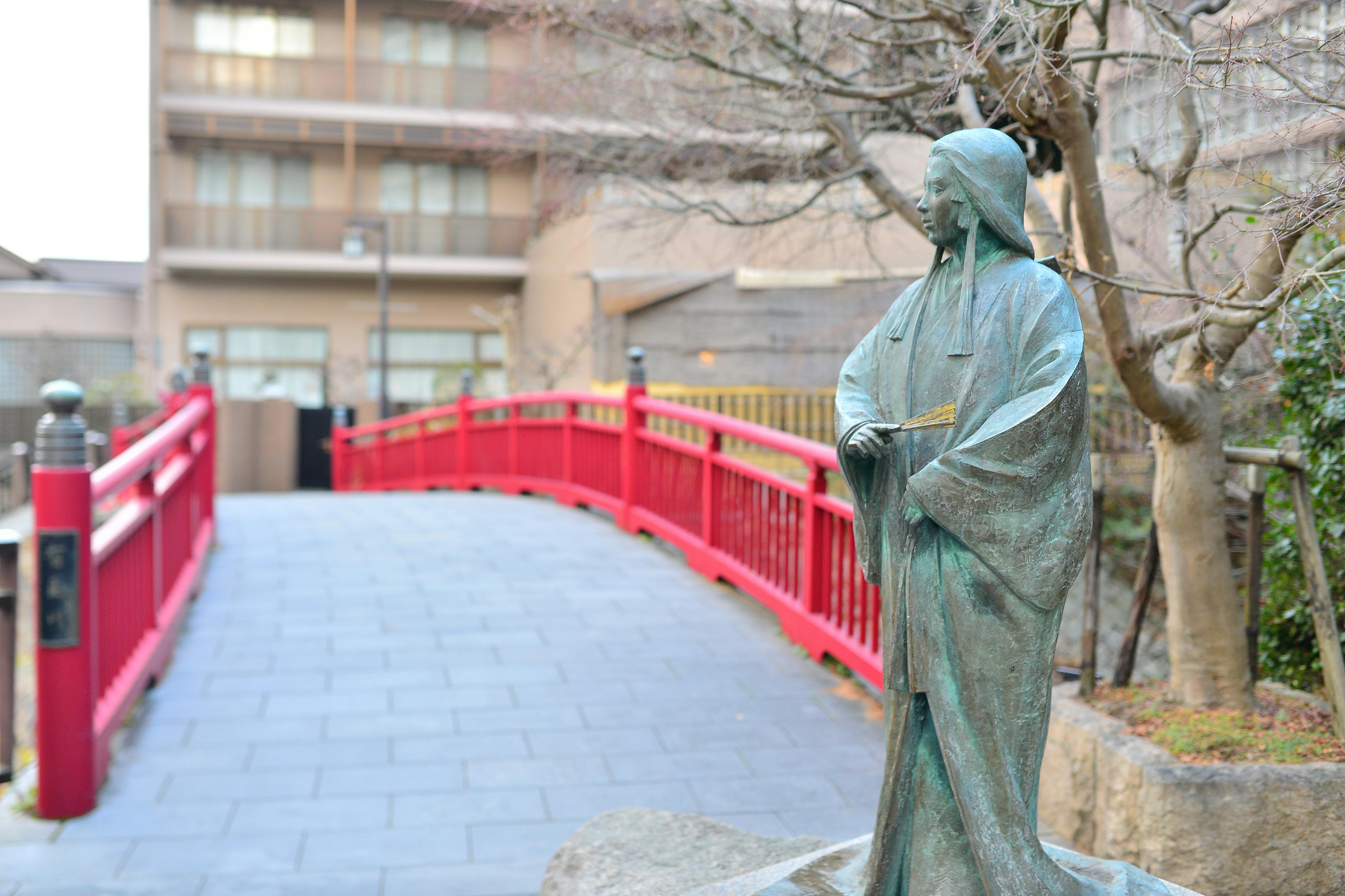 A bronze statue of a figure in traditional Japanese clothing stands near a red arched bridge, with leafless trees and modern buildings in the background.