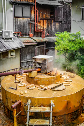 A large, round, yellow industrial vat emits steam from a central metal box, surrounded by several circular lids. The background shows weathered wooden buildings, pipes, and a leafy green tree.