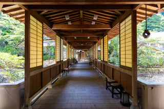 A covered wooden walkway with shoji-style panel walls and a tiled floor, lined with benches, extends into a lush green garden under natural daylight.