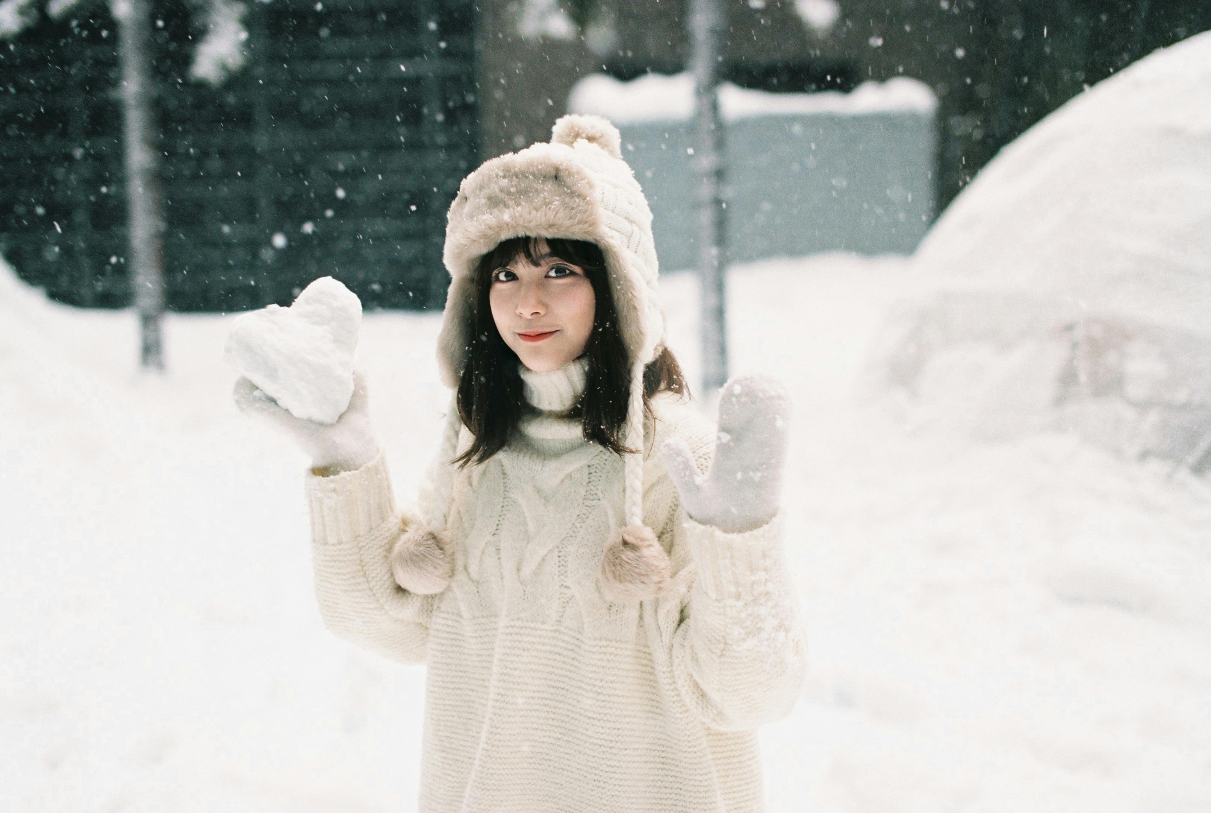 A young woman in a white sweater, hat, and mittens stands outside in falling snow, holding a heart-shaped snowball and smiling. Snow covers the ground and background.