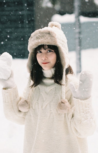 Skiing A young woman in a white sweater, hat, and mittens stands outside in falling snow, holding a heart-shaped snowball and smiling. Snow covers the ground and background.