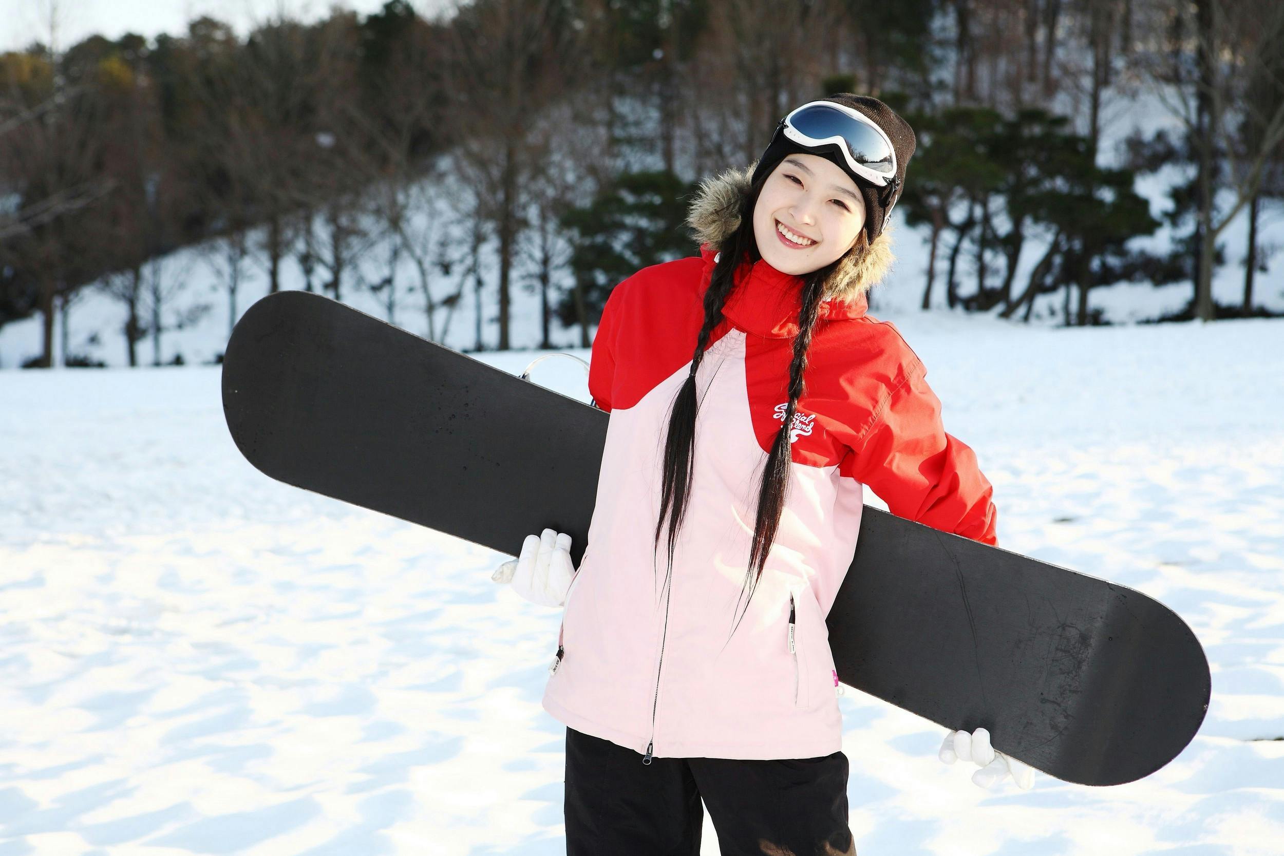 A smiling person in a red and pink jacket, black pants, and ski goggles stands in the snow holding a snowboard, with snow-covered ground and trees in the background.