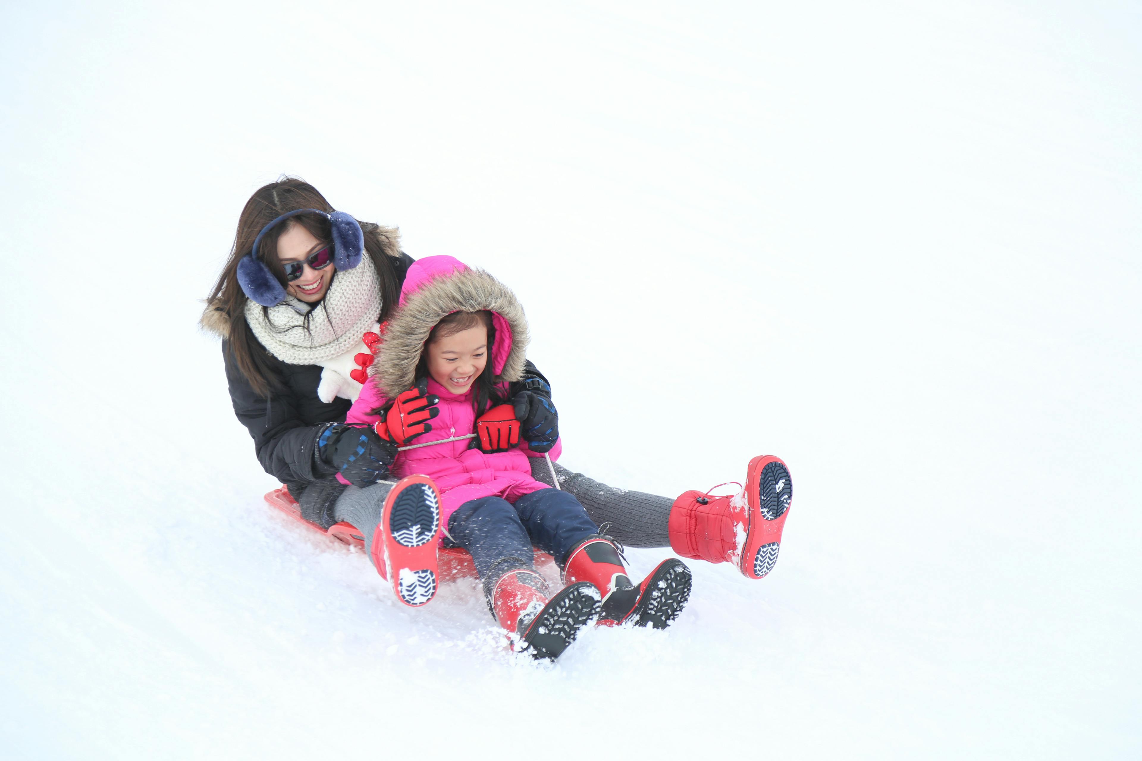 A woman and a young girl, both bundled in winter clothes, slide down a snowy hill together on a red sled, smiling and enjoying the ride. The background is bright white with fresh snow.