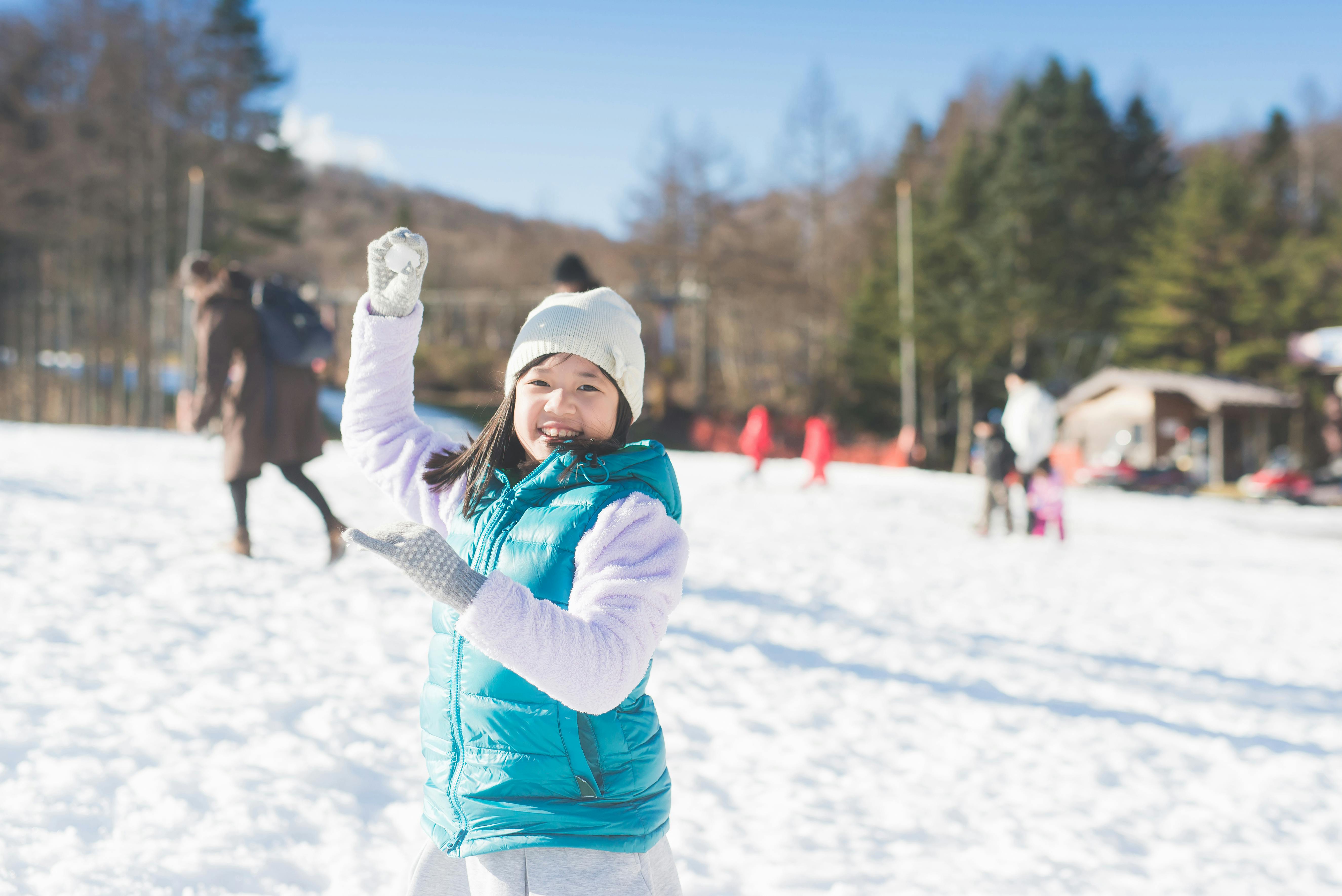A smiling child in a blue vest and white hat stands in a snowy field, holding a snowball and ready to throw it. People and trees are visible in the bright, sunlit background.