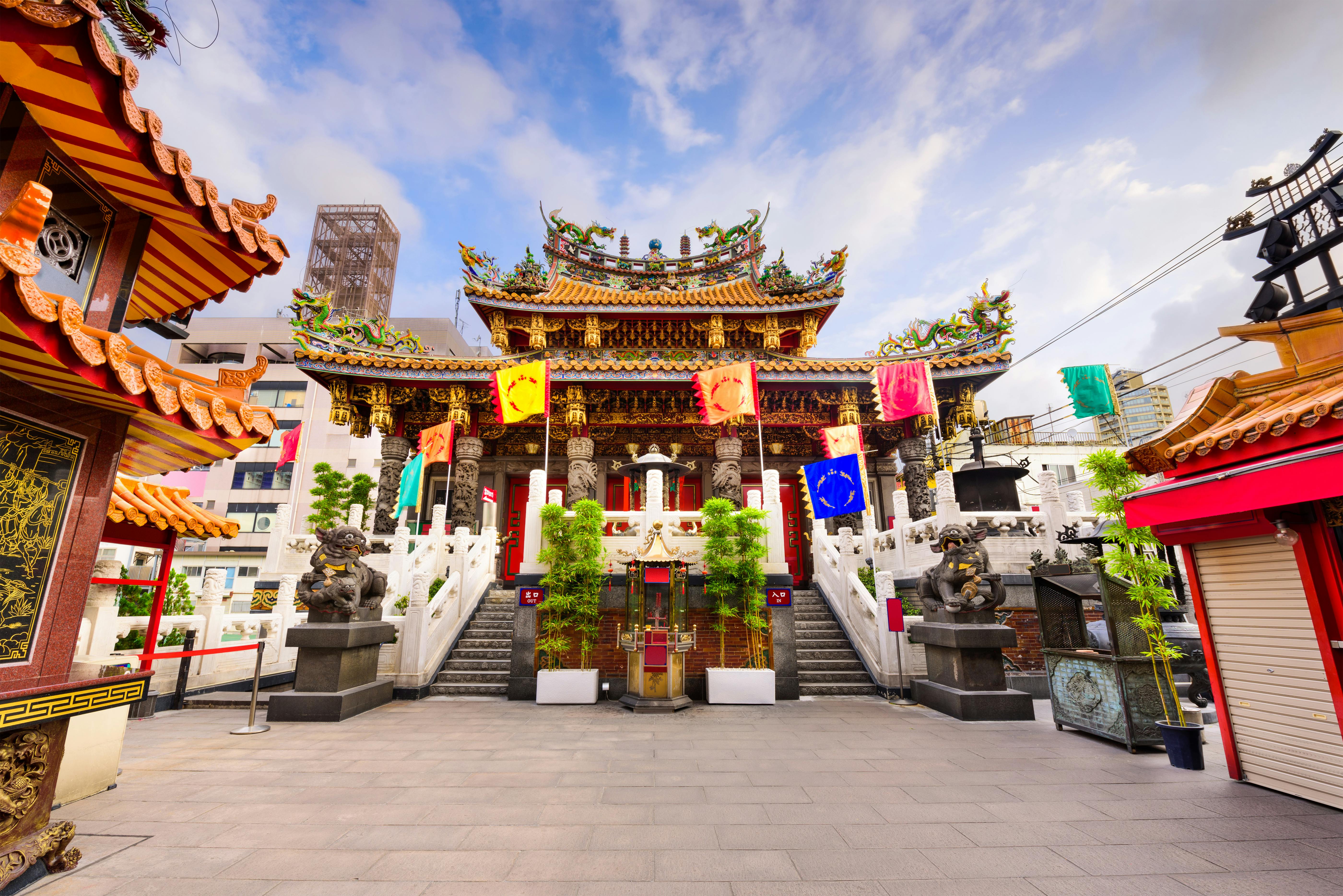 A traditional Chinese temple with ornate, colorful roof decorations, multiple flags, stone lion statues, white staircases, and green plants, set against a backdrop of modern city buildings and a partly cloudy sky.