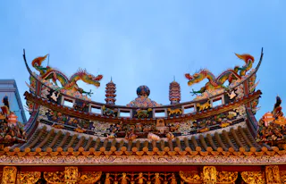 Ornate temple rooftop with intricate carvings, colorful details, and two dragon statues facing each other against a blue sky. The design is traditional East Asian architecture.