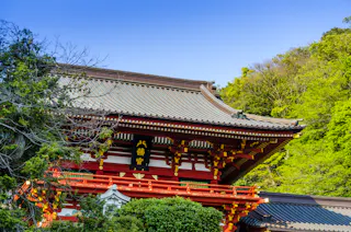 A traditional Japanese temple with a tiled roof and red, white, and gold accents stands among lush green trees under a clear blue sky.