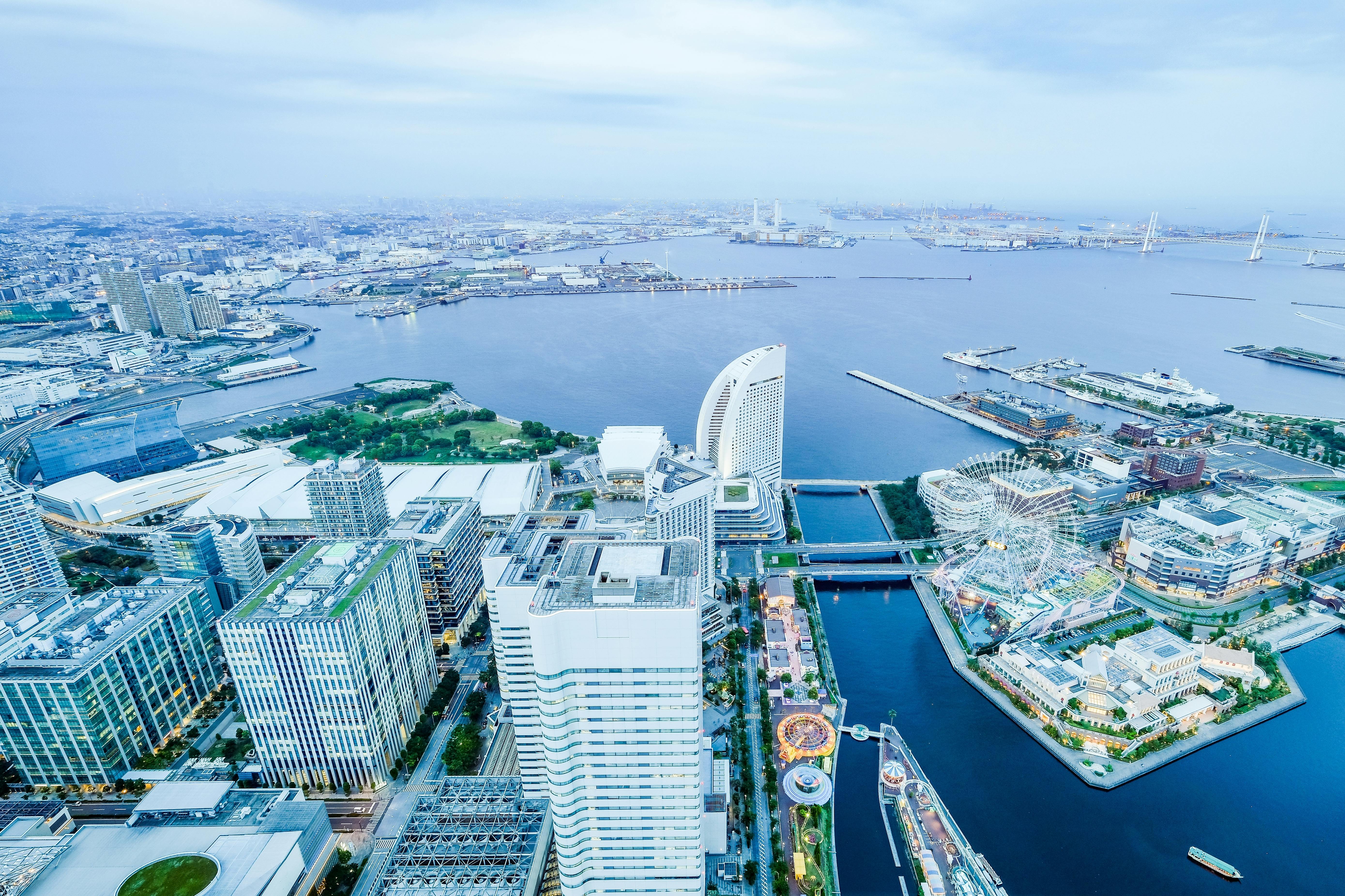 Aerial view of a modern city with tall buildings, waterfront parks, a curved skyscraper, and a bay with docks and ships, under a cloudy sky.