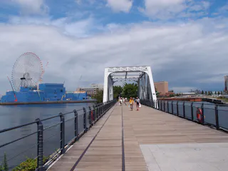 A wide pedestrian bridge with people walking, spanning over water towards a cityscape. A large Ferris wheel and blue building are visible on the left under a partly cloudy sky.