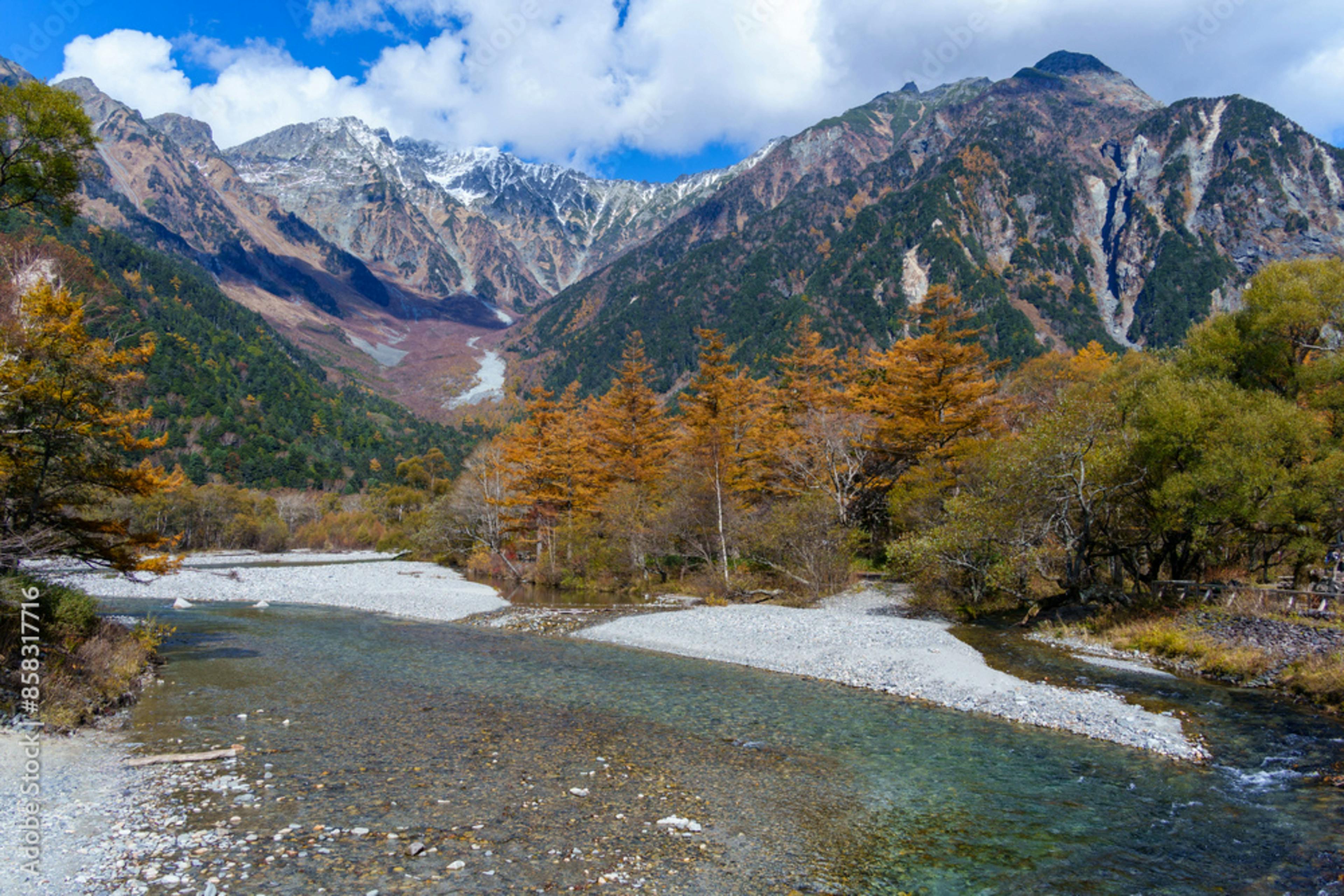 A clear river winds through a rocky bed surrounded by autumn trees with orange and yellow leaves, set against tall, rugged mountains with patches of snow under a blue sky with scattered clouds.