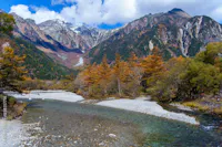 A clear river winds through a rocky bed surrounded by autumn trees with orange and yellow leaves, set against tall, rugged mountains with patches of snow under a blue sky with scattered clouds.