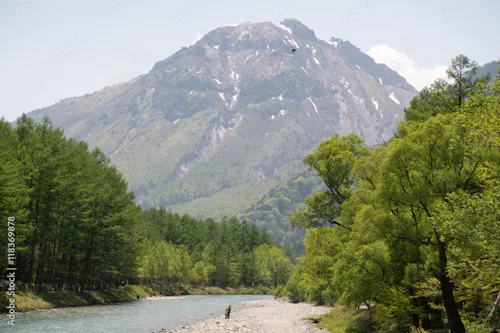 A person stands on a rocky riverbank surrounded by lush green trees, with a clear river flowing beside them. In the background, a tall, forested mountain with patches of snow rises against the sky.