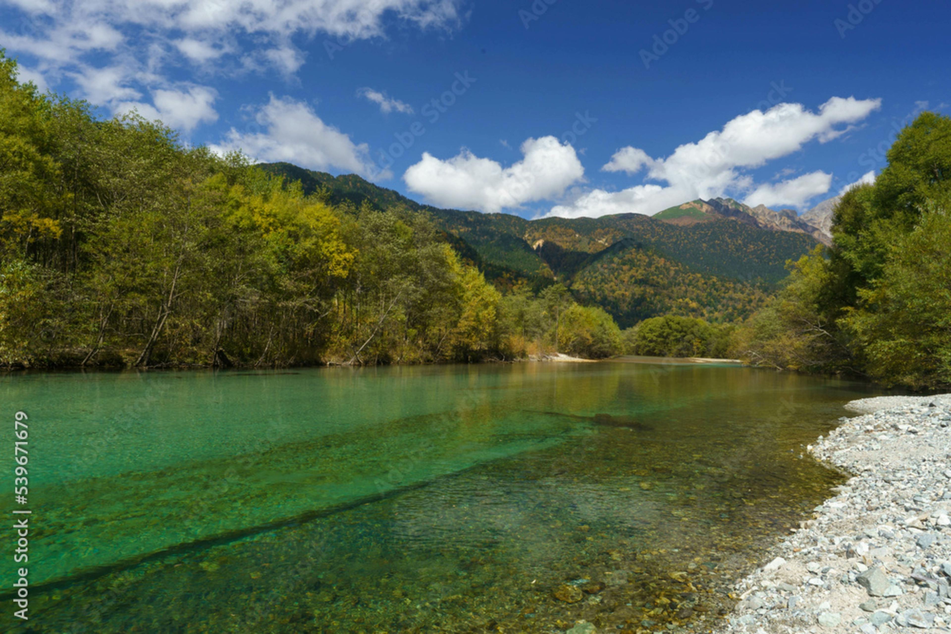 A clear, green river flows gently between tree-lined banks, with pebbles visible underwater. Forested mountains rise in the background beneath a blue sky with scattered white clouds.
