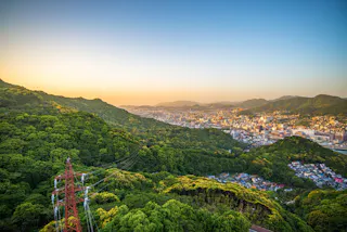 A city nestled between lush green hills under a clear blue sky at sunset, with a red electricity pylon and power lines in the foreground.