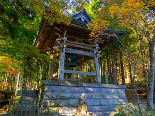 A traditional Japanese bell tower stands among autumn trees, surrounded by vibrant yellow and green foliage. A small stone statue sits on the stone base in front of the structure, with sunlight filtering through the leaves.