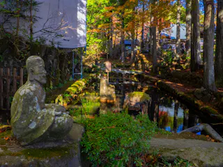 A stone statue sits by a tranquil pond surrounded by trees with autumn foliage, reflecting in the water. Sunlight filters through the leaves, creating a peaceful and serene atmosphere in the garden.
