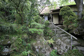 A traditional Japanese garden with lush greenery, stone steps, a wooden handrail, and a small house partially hidden among trees and shrubs.