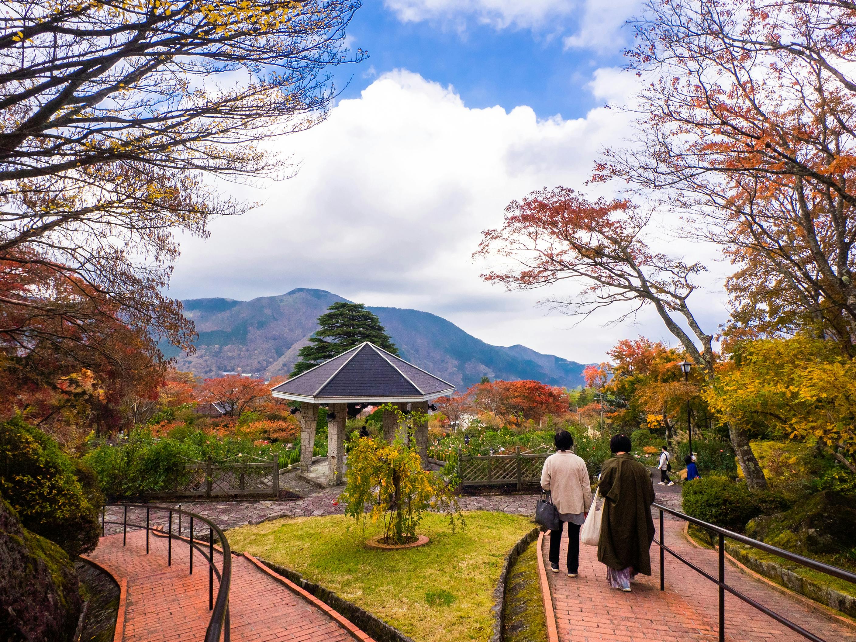 Two people walk along a brick path in a park with autumn foliage. A pavilion stands in the background against a backdrop of mountains and a partly cloudy sky. Bare trees and colorful leaves surround the scene, creating a peaceful atmosphere.
