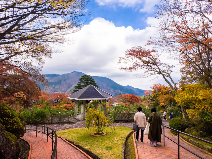 Two people walk along a brick path in a park with autumn foliage. A pavilion stands in the background against a backdrop of mountains and a partly cloudy sky. Bare trees and colorful leaves surround the scene, creating a peaceful atmosphere.