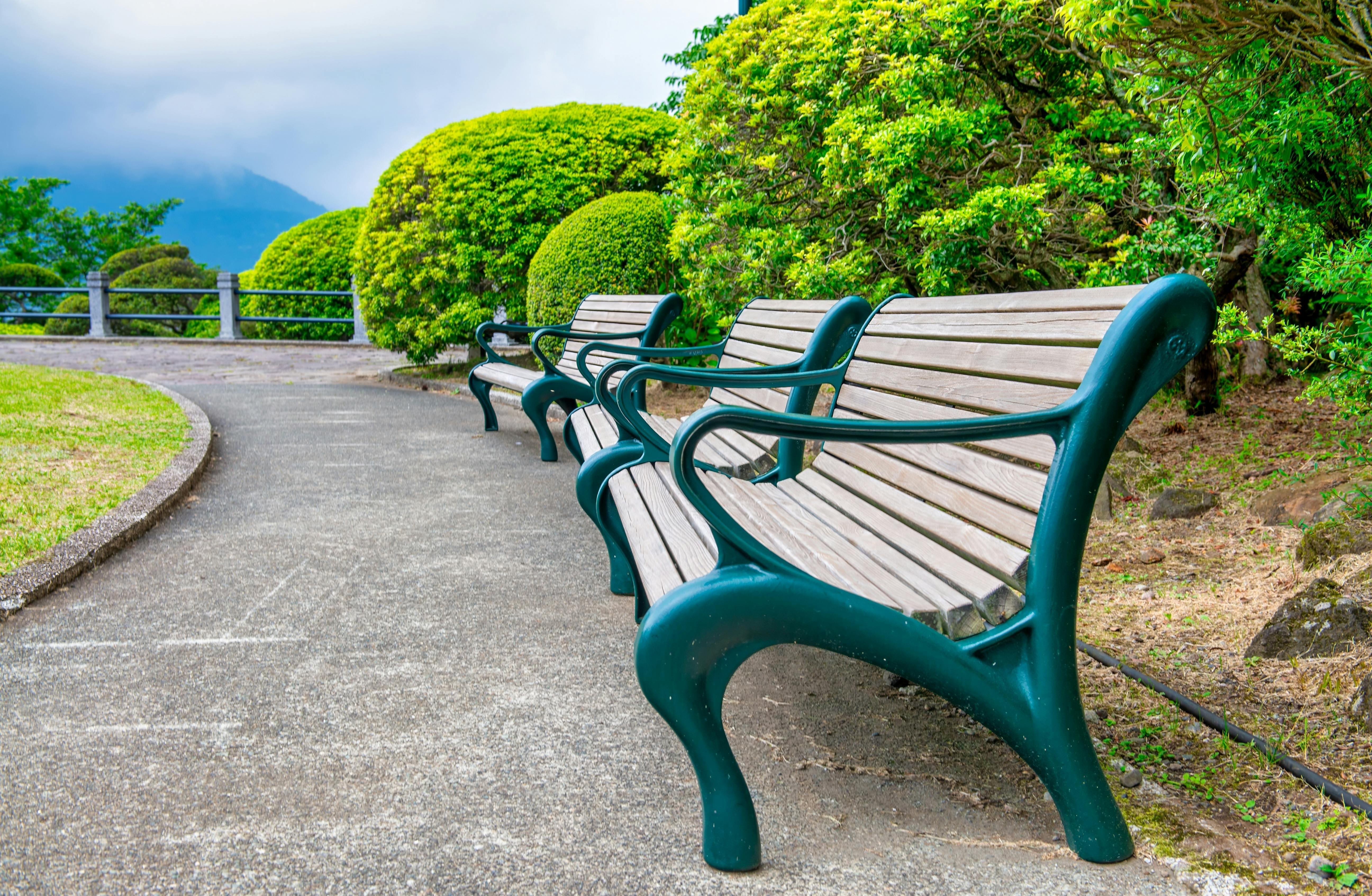 A row of empty wooden benches with green metal frames lines a curved pathway in a lush garden, surrounded by neatly trimmed bushes and trees.
